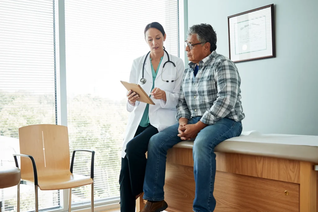 GP with a tablet talking to a patient seated on an examination couch in a consultation room