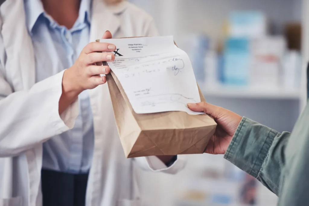 Pharmacist handing a paper prescription bag to a patient across the pharmacy counter